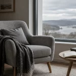 Sillón vacío junto a una ventana gris y una mesa con libros y un cuaderno.